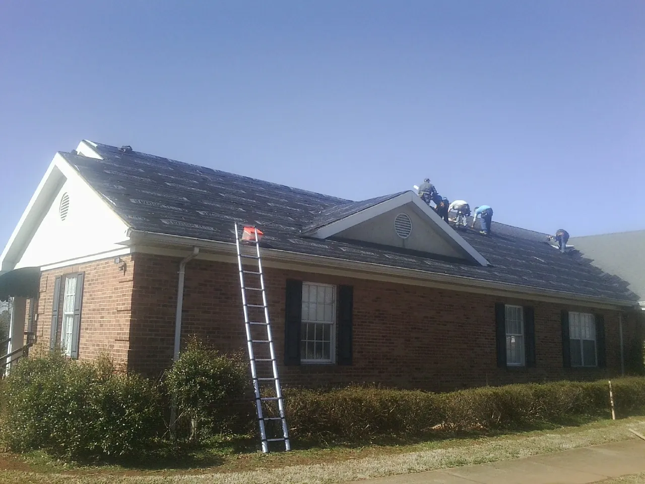Crew tearing shingles off a brick ranch roof with a ladder set — Emergency Roof Repair work by our crew in Crane